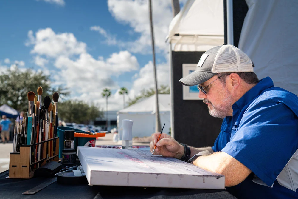 An artist painting outdoors at the Space Coast Art Festival, with brushes and supplies on display.